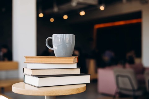 A serene coffee shop scene with a stack of books and a gray mug on a wooden table, set against a