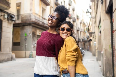 Two friends, one wearing sunglasses and both dressed in colorful sweaters, share a joyful moment on