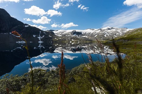 A crystal-clear mountain lake in Norway reflects snow-dusted peaks, bright skies, and lush greenery