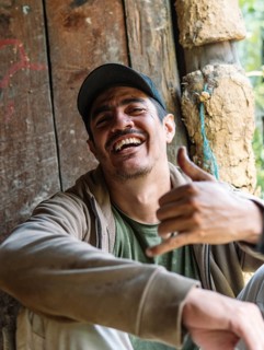 A cheerful man wearing a cap and jacket laughs joyfully while seated in a rustic wooden structure