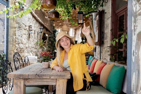 A young, cheerful woman in a yellow shirt sits at a cafe table, wearing a straw hat. She is smiling