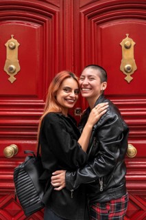 A joyful lesbian couple embraces lovingly in front of striking red doors with golden details. The