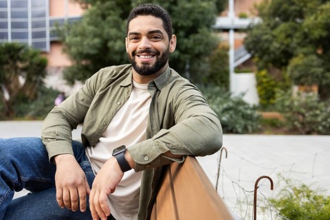 A man in a casual shirt and jeans sits on a bench of the city, smiling warmly at the camera. The