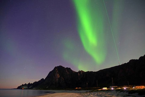Northern lights glow over a rocky coastline with beach and illuminated areas at night, Laser,