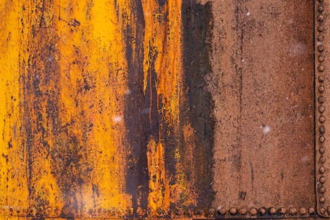 Rusted steel wall with peeling orange paint and rows of rivets, detailed view of an industrial
