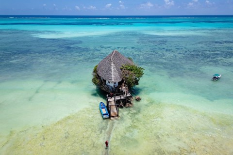 Aerial view of the building surrounded by turquoise water and white sand beach with boats under a