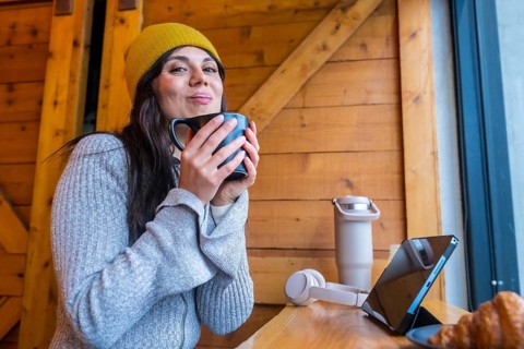 Woman in a yellow beanie staying cozy enjoying a cup of coffee while working remotely on a digital