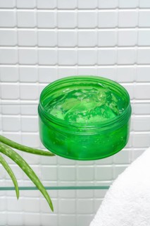 Jar of aloe vera gel next to fresh aloe leaves and a white towel in a bright bathroom setting