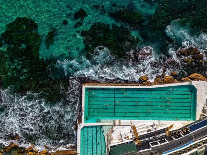 Stunning aerial shot of a seaside pool next to vibrant turquoise ocean, with swimmers enjoying the