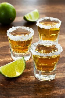 Three glasses with tequila and salt on the rims are arranged on a wooden table. Each glass has a