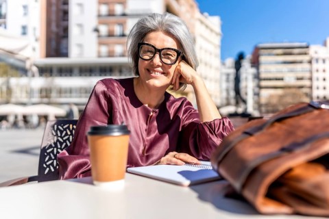 Grey haired woman at an outdoor cafe smiling at camera, enjoying sunny city day with coffee,