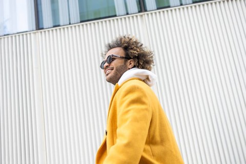 A confident afro man in a bright yellow coat and sunglasses smiles warmly, expressing positivity