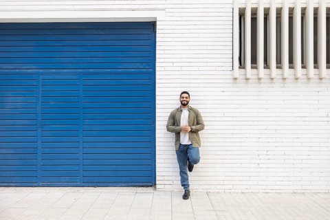 A man stands casually against a white brick wall next to a blue garage door in the city. His