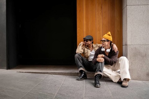 A cheerful gay couple sits together on an urban street outside a wooden and stone wall. They are