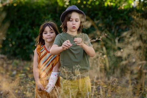 Two children are joyfully playing in a sunlit field. One is wearing a hat and is curious about a