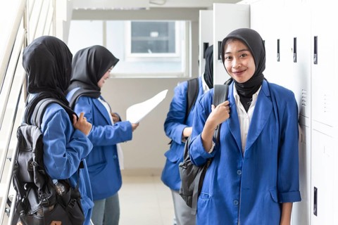 Asian Muslim students in blue blazers and hijabs stand and chat in a school corridor. They carry