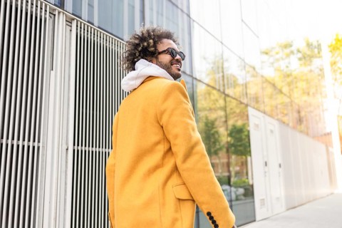 A stylish afro man in a vibrant yellow coat and sunglasses walks confidently past a modern glass