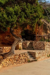 Stone stairs leading to a small cave in the rock face, shaded by a juniper tree, in puerto cristo,
