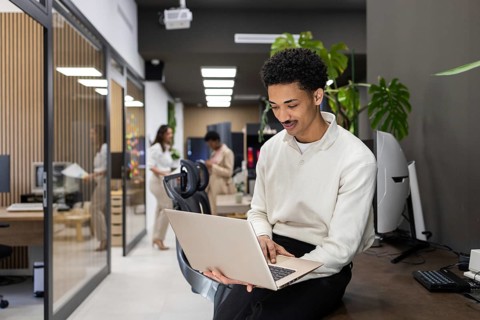 A professional office environment with a man using a laptop while sitting on a desk, and colleagues