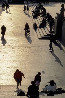 A group of people are walking along the street, some are sitting on the steps. The scene is casual