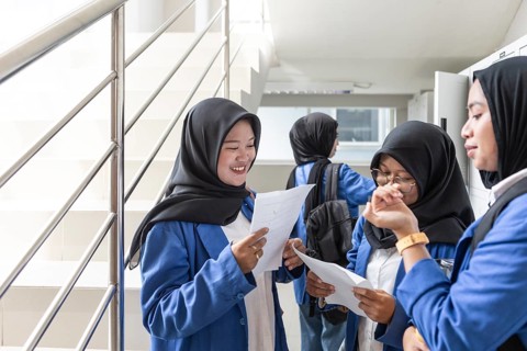 Asian Muslim students in hijabs engage in a lively discussion, holding papers, as they prepare for