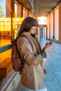 Young woman student wearing a backpack walking and smiling while checking her mobile phone in a