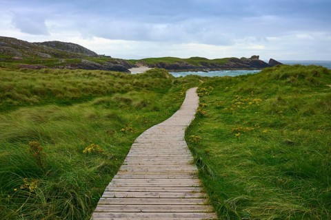 A winding wooden path leads through grassy dunes to a distant sea under overcast sky, Clachtoll,