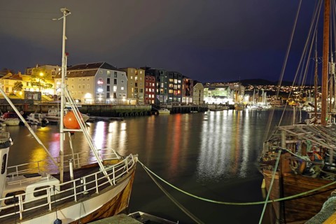 Boats in illuminated nocturnal harbor next to colorful buildings and calm water, Nidelva,