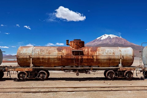 Abandoned rusty tank truck in a desert landscape with mountains in the background, railroad cars in