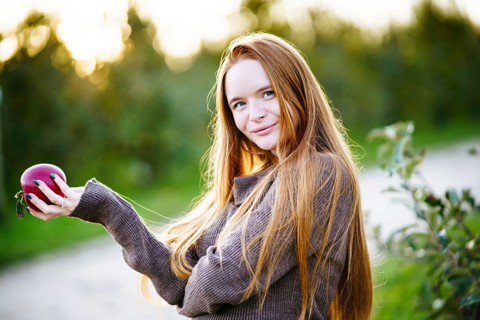 A radiant pregnant woman with long red hair stands outdoors, gently holding a red apple. Soft