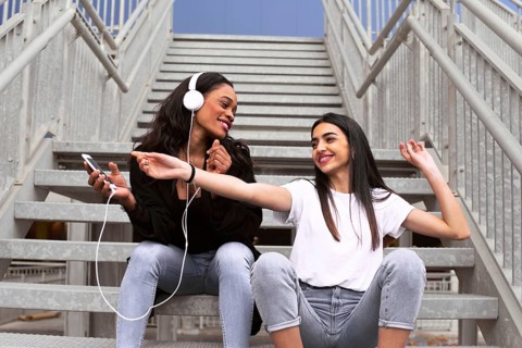 Two friends sit on outdoor stairs, sharing earphones and music from a smartphone. They appear