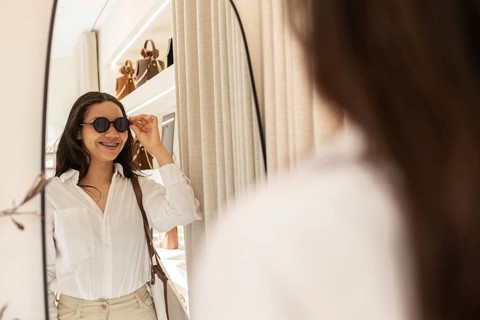 A stylish woman tries on sunglasses and smiles in front of a mirror inside a chic bag store. The