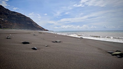 A secluded black sandy beach, volcanic sand with scattered stones under a wide sky, hiking,