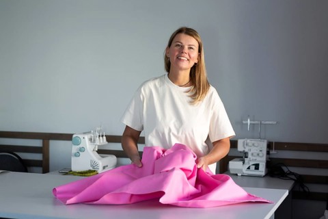A woman stands confidently in a sewing workshop, holding vibrant pink fabric. She is surrounded by