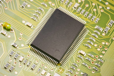 A detailed view of a microchip positioned on a green circuit board. The intricate paths and