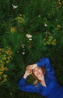 A cheerful blonde woman lies joyfully in a grassy field, surrounded by vibrant green foliage and