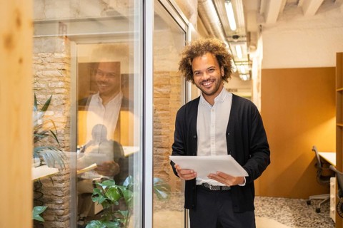 A smiling man with a tablet stands in a modern coworking space, showcasing a bright, collaborative