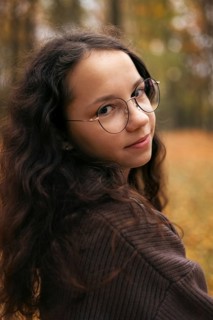 A teenage girl with long curly brown hair and glasses looks over her shoulder in an autumn forest.