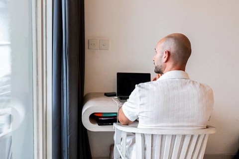 A man seated at a desk in a hotel room, working on a laptop. The bright interior and modern