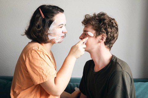 A young man and woman enjoy a playful skincare routine together in their living room. Both are