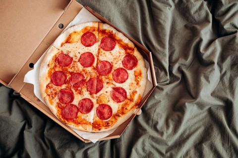 From above, a freshly baked pepperoni pizza sits in its box, placed on a rumpled grey bed sheet,