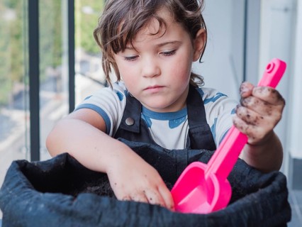 A focused young girl is seen planting a sunflower seed in a dark planter with a pink shovel, her