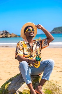 Young man wearing sunglasses and straw hat holding a pineapple shaped cocktail while relaxing on a