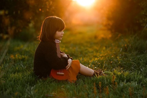 A young child sits in a lush green field with orange trees holding a ukulele, bathed in the warm