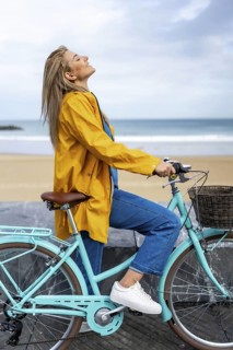 Young woman wearing yellow raincoat enjoying fresh air while riding bicycle on a cloudy day by the