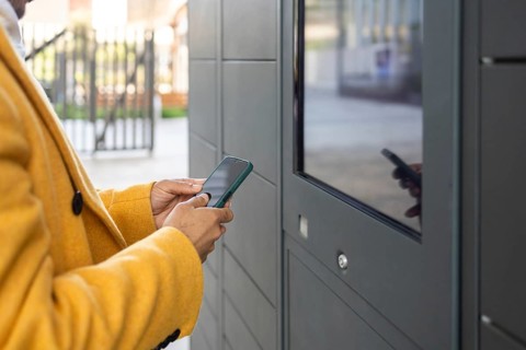 A businessman in a yellow coat uses a smartphone at an outdoor digital kiosk. The setting is urban