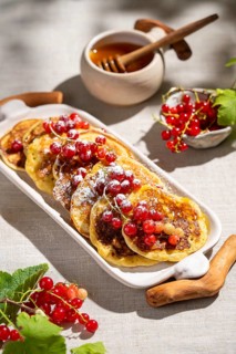 From above, a white platter of golden pancakes topped with vibrant red currants and dusted with