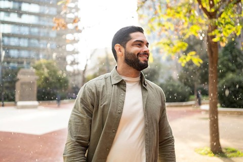 A man with a beard smiles while enjoying a light rain in a park of the city. He wears a light