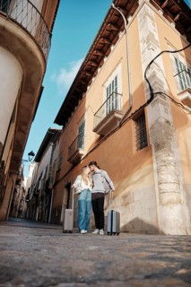 A young couple strolls with luggage in a charming Majorcan street, searching for their dream