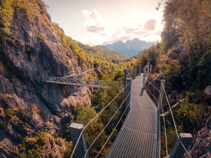 A metal bridge spans a scenic mountain gorge in the Pyrenees, surrounded by lush greenery and rocky